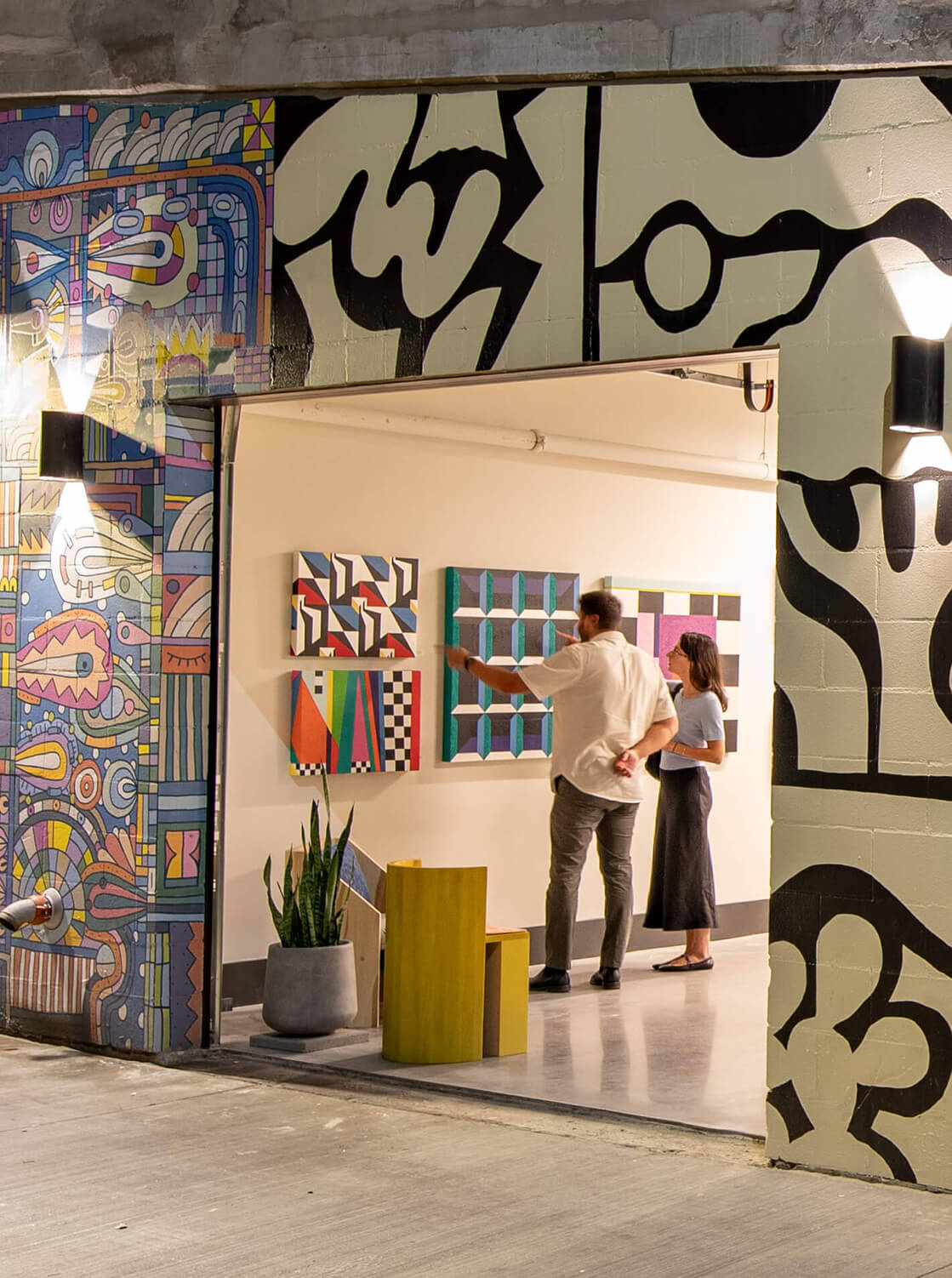 Two people viewing colorful abstract artwork inside a gallery, framed by vibrant mural-covered walls.