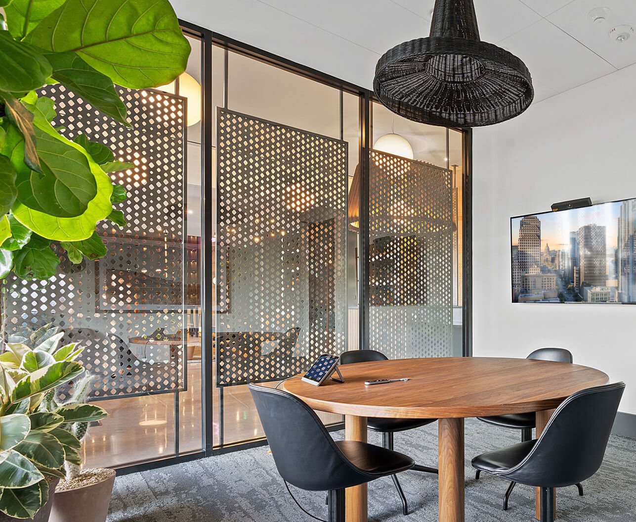 Modern meeting room with a round wooden table, black chairs, a TV displaying a cityscape, and decorative perforated panels.