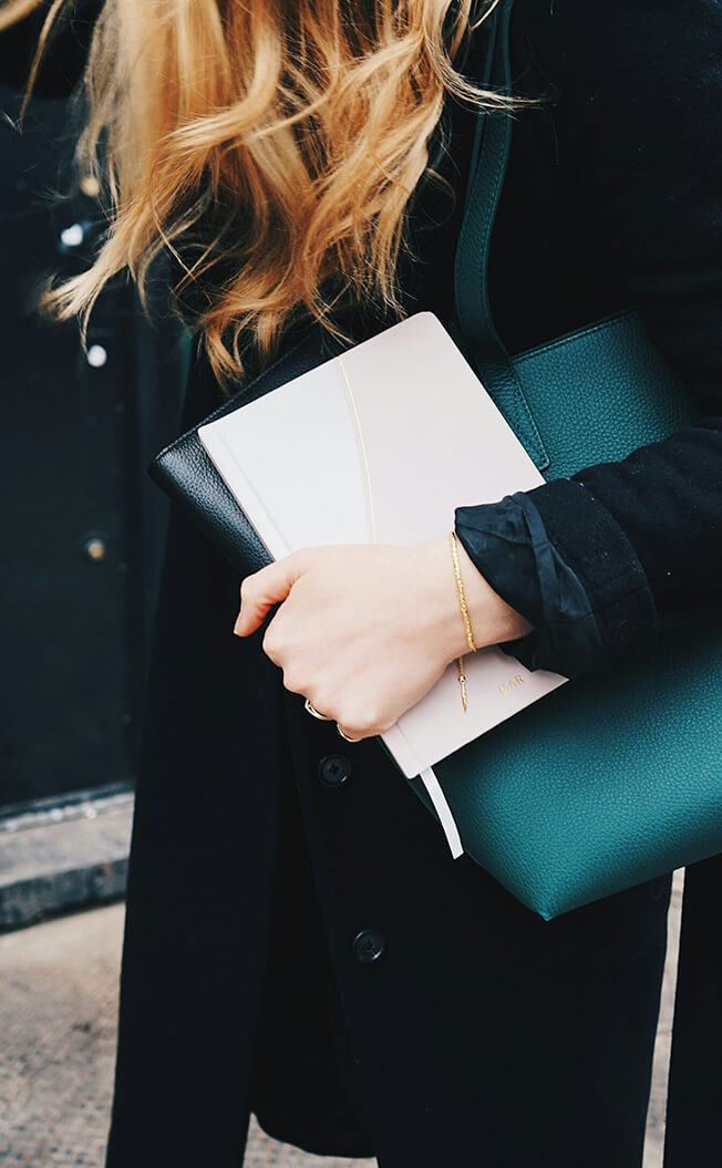 Close-up of a woman holding notebooks and a green tote bag, wearing a black coat and a delicate gold bracelet.