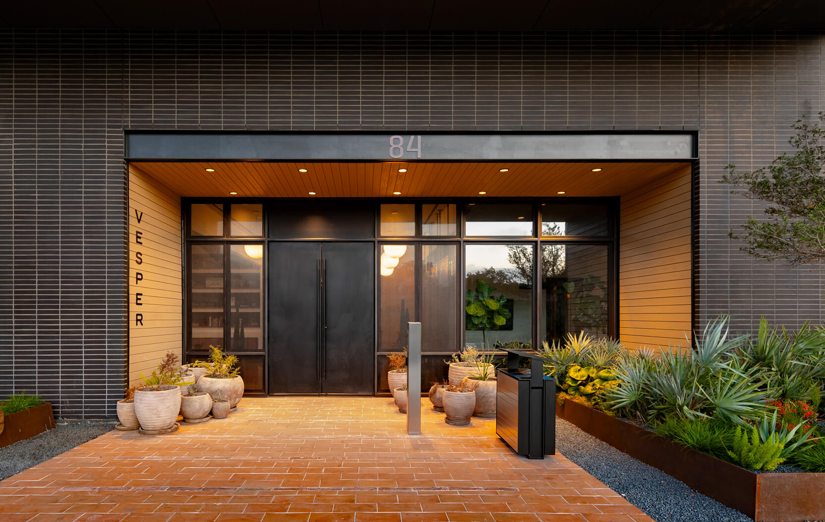 Elegant building entrance with black doors, terracotta tile flooring, potted plants, and modern landscaping.