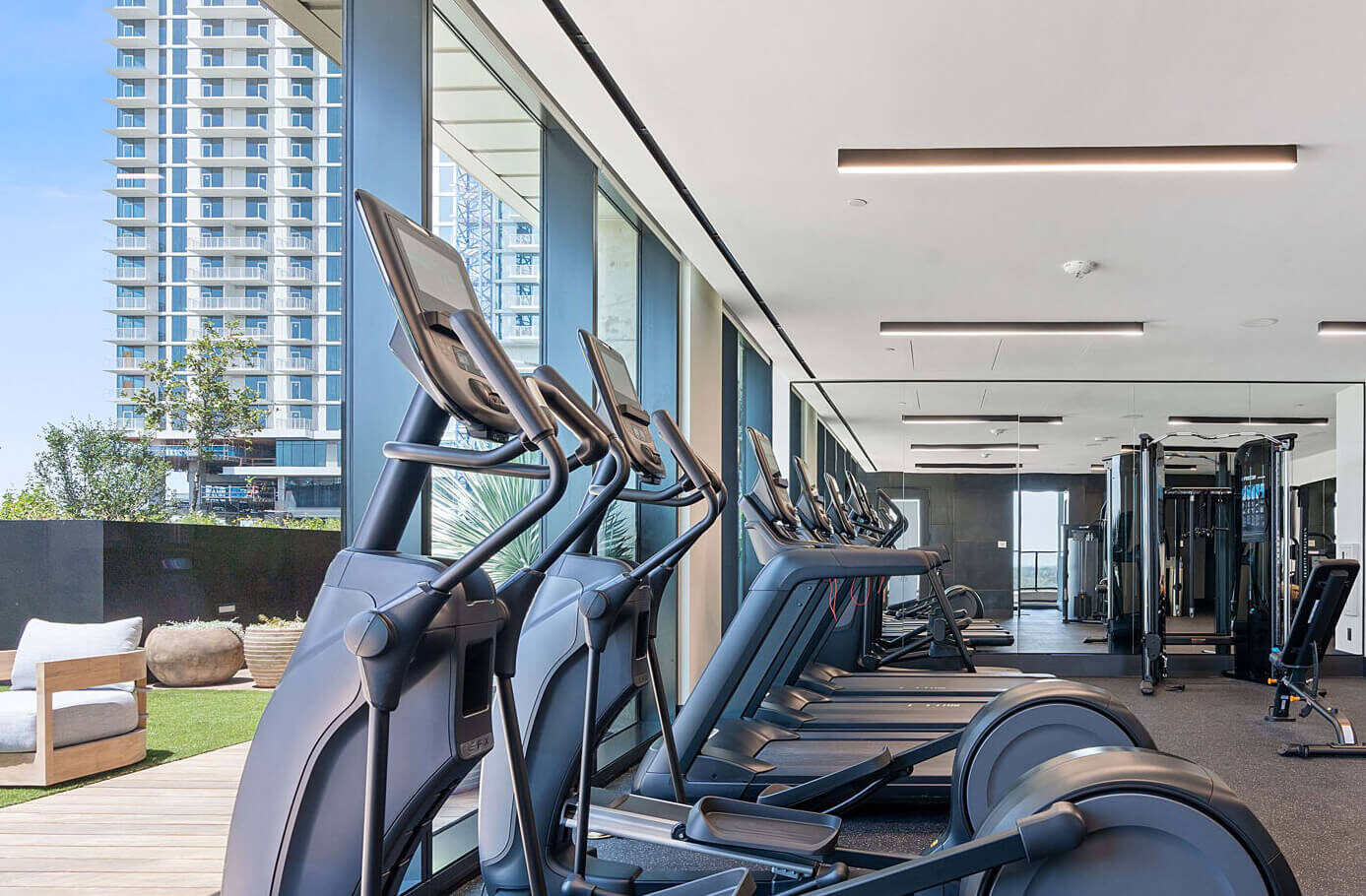 Modern fitness center with treadmills, ellipticals, weight machines, and floor-to-ceiling windows overlooking a cityscape.