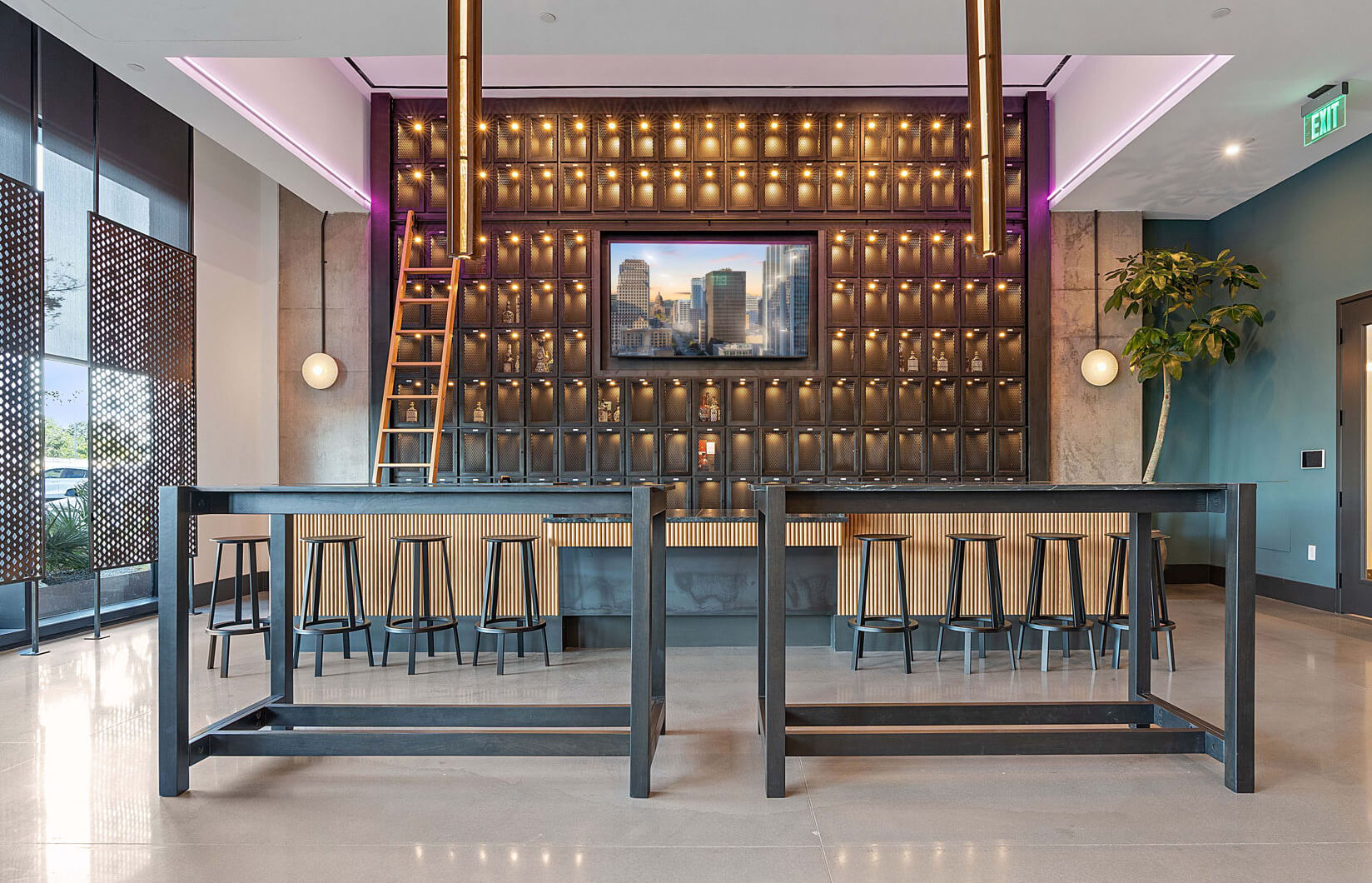 Modern bar area with a black metal liquor lockers in the lobby of Vesper Residences, illuminated bottles, a wooden ladder, barstools, and a cityscape display screen.