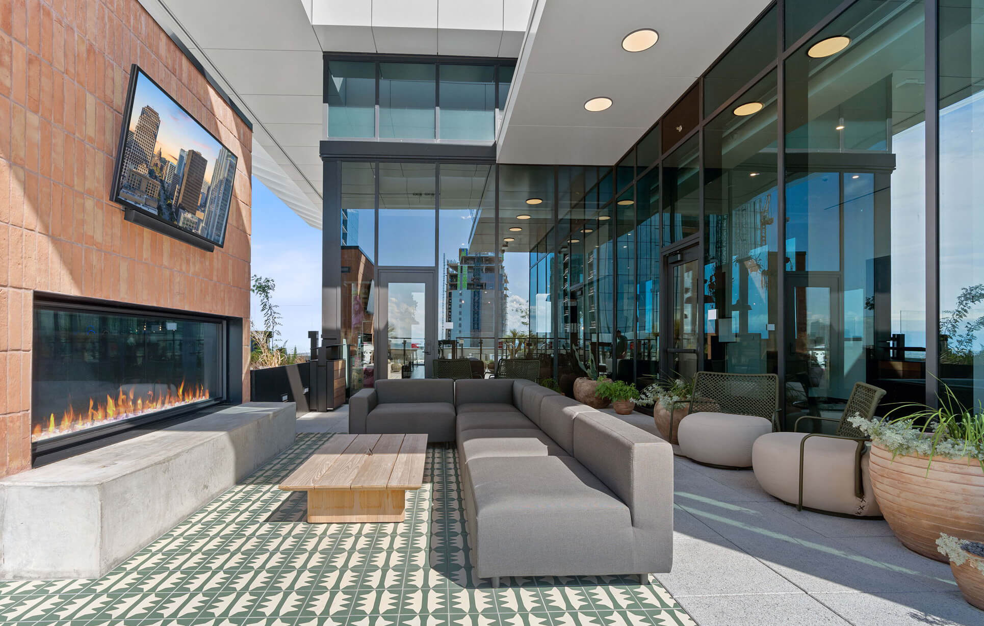 Modern outdoor lounge with a gray sectional, a fireplace, potted plants, tiled flooring, and a mounted TV overlooking the city.