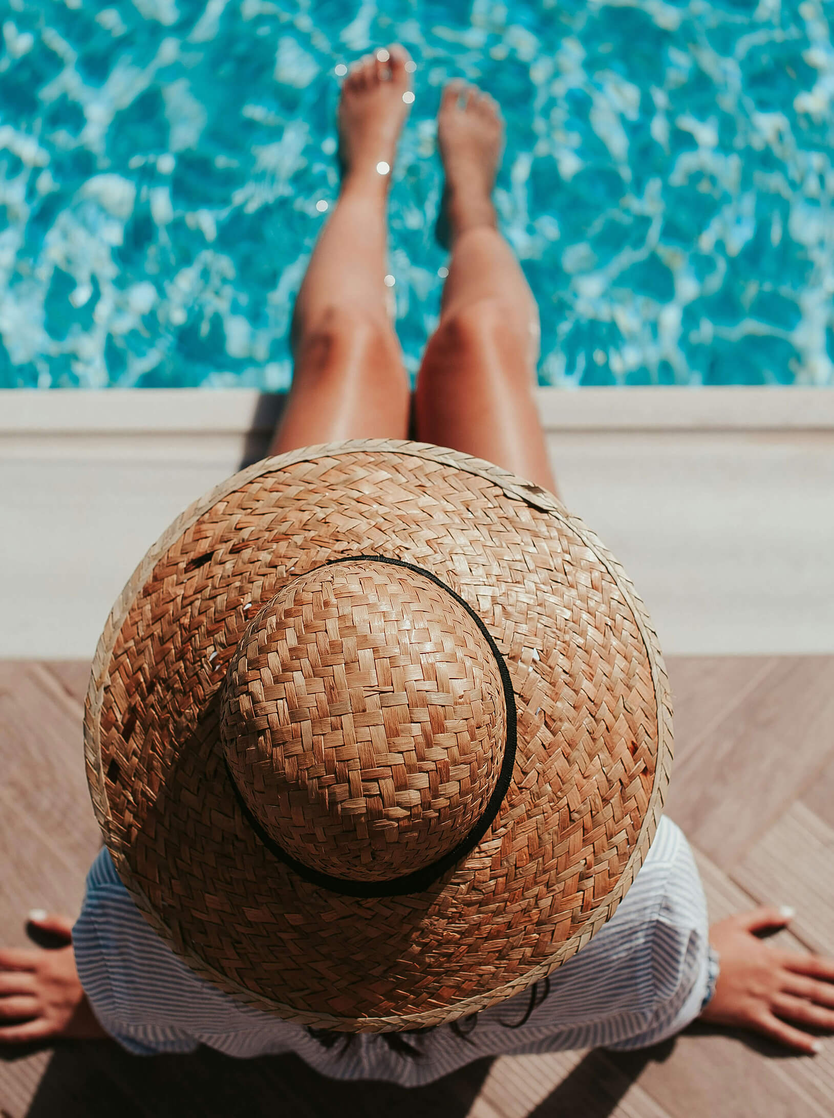 Person wearing a straw hat, sitting by a pool with legs outstretched, enjoying the bright blue water.