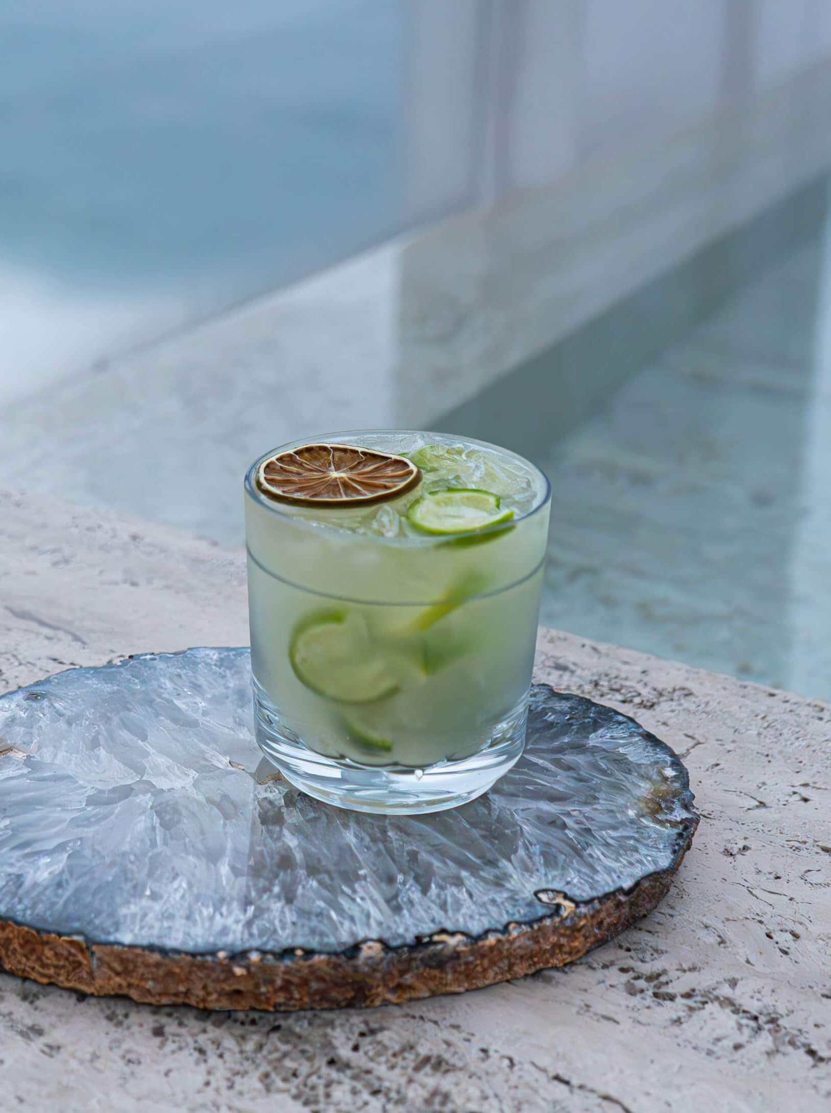 Refreshing cocktail with lime slices and ice, served on a decorative crystal coaster near a pool.