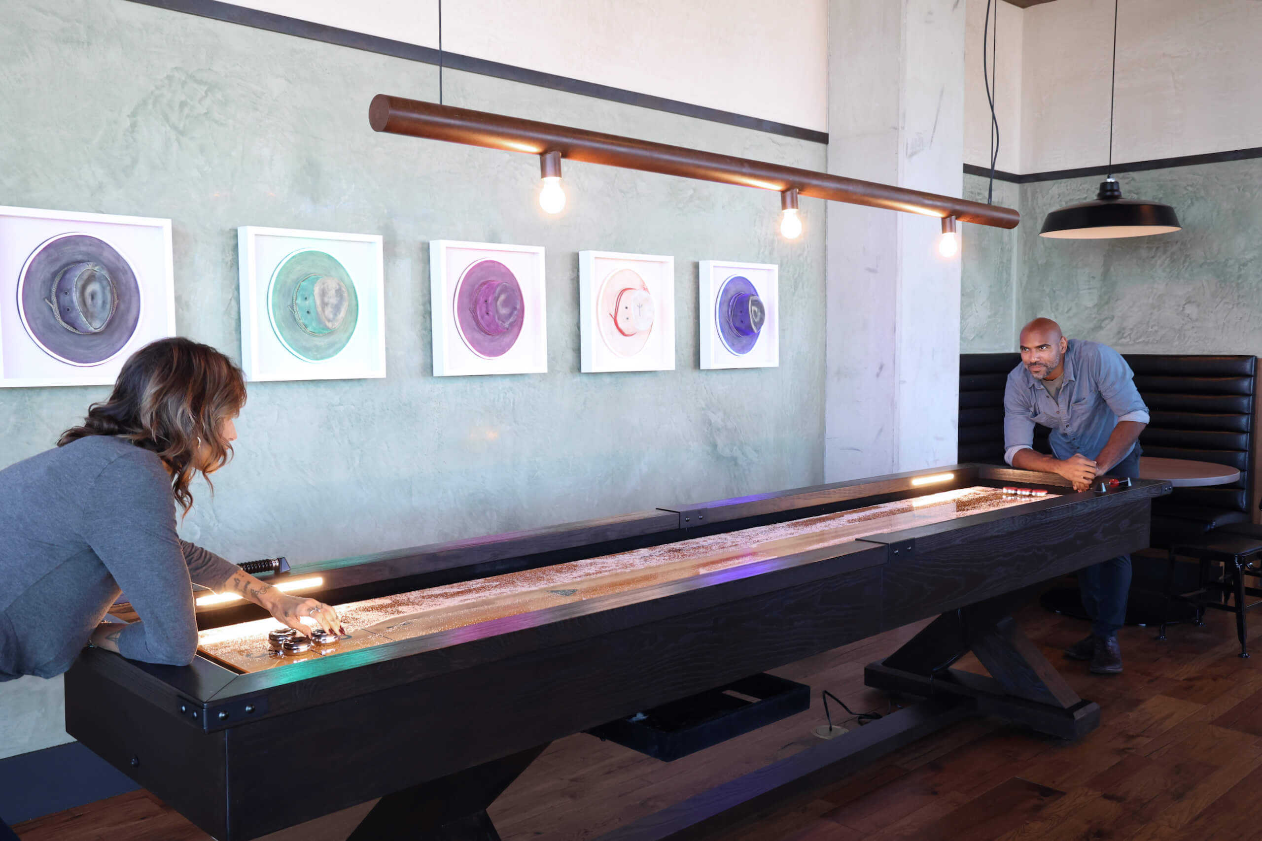 Two people playing shuffleboard in a modern game room with colorful artwork, pendant lighting, and a leather booth.