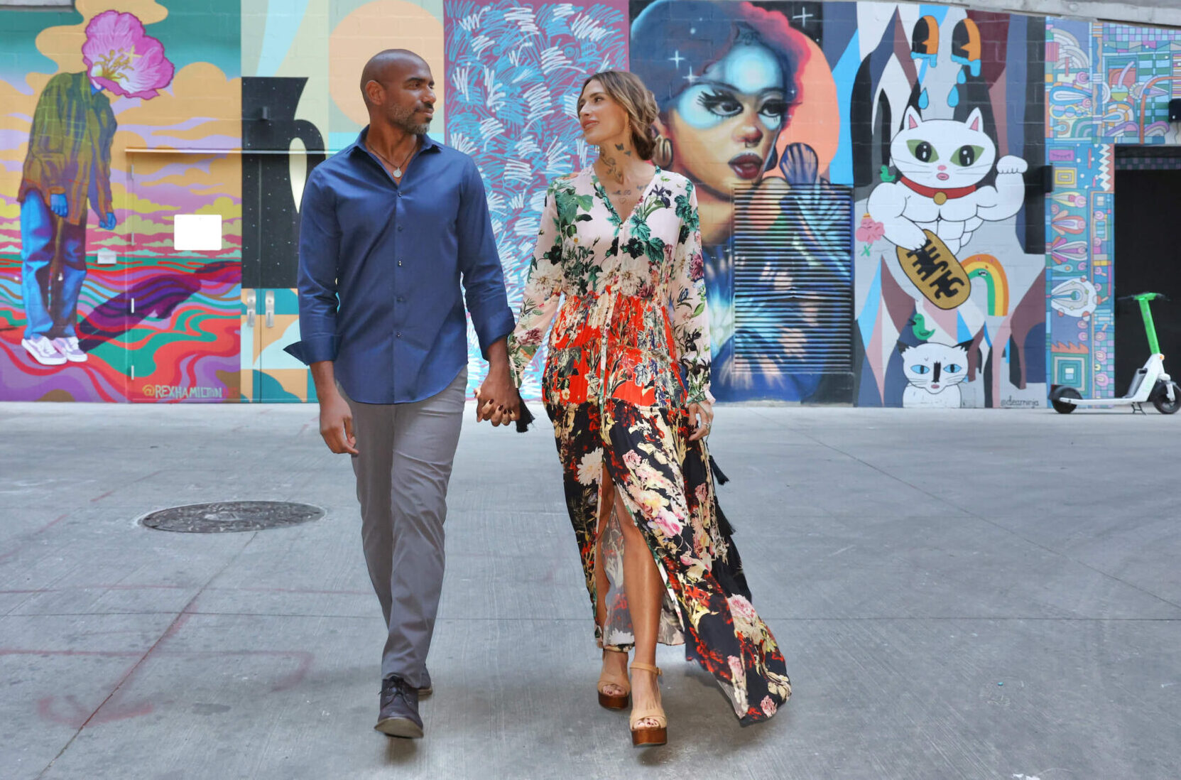 Couple walking hand in hand in front of vibrant street art murals, with the woman in a floral dress and the man in a blue shirt.