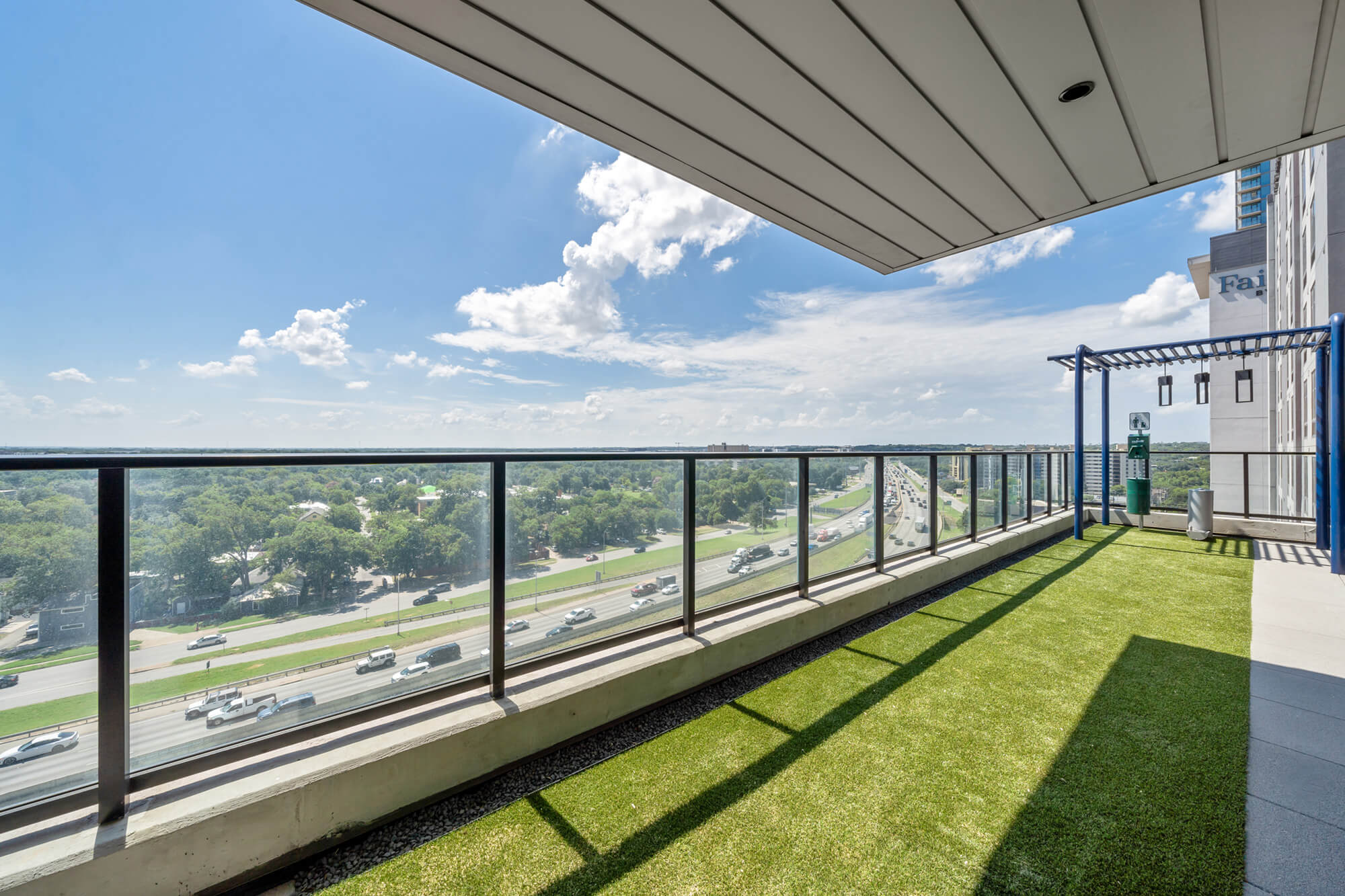 Spacious balcony with glass railings, artificial turf, a pergola, and scenic views of a highway and greenery under a bright sky.