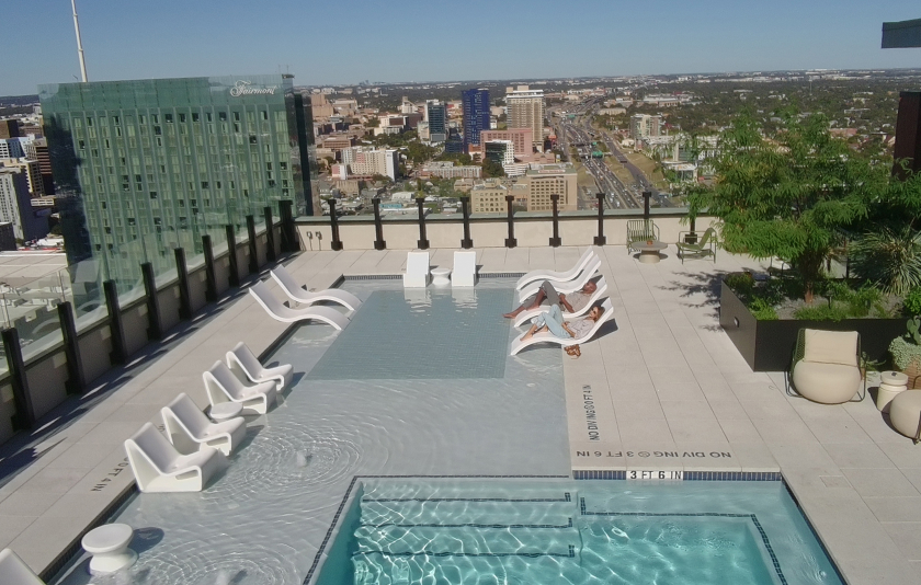 Rooftop pool with lounge chairs, shallow water seating, and panoramic views of a city skyline and greenery.