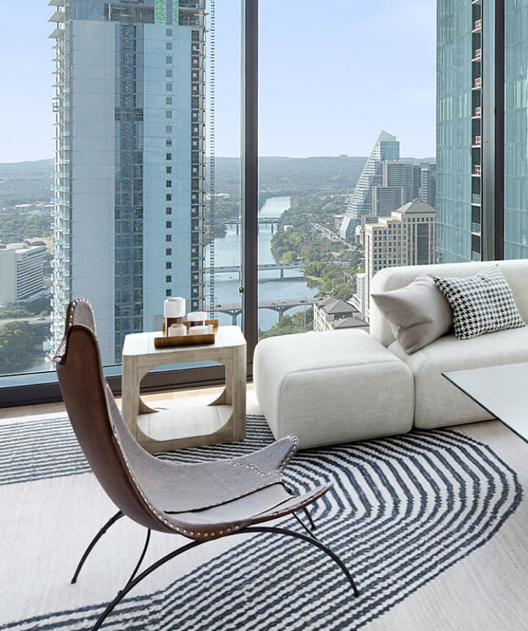 Image of a light filled living room with floor to ceiling windows that look out upon the Austin skyline and Lady Bird Lake. There is a modern chair in focus with a rug that has a curved line pattern.